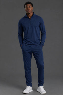 A man modeling a matching navy blue quarter-zip pullover and jogger pants set, paired with white sneakers, against a solid dark gray studio background. || Navy