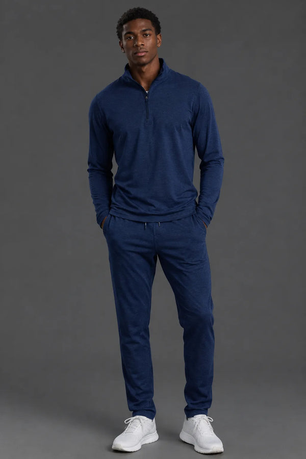 A man modeling a matching navy blue quarter-zip pullover and jogger pants set, paired with white sneakers, against a solid dark gray studio background. || Navy