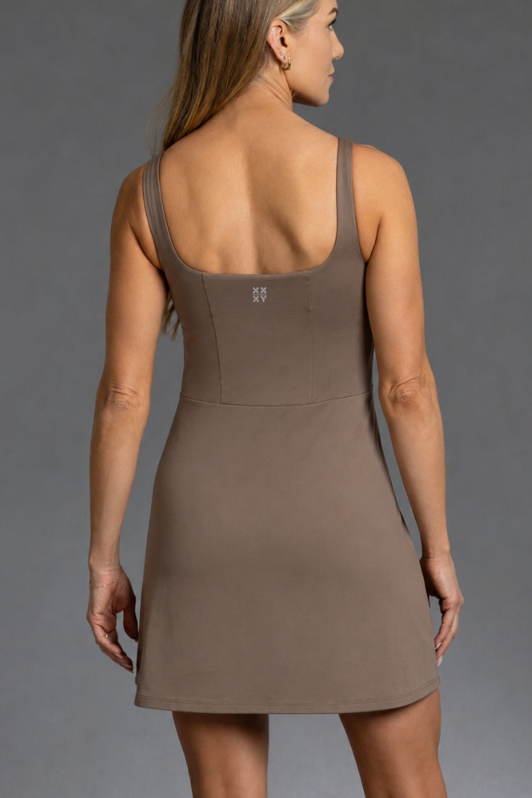 A woman shown from the back wearing a sleeveless, scoop-neck mini dress in a solid taupe-brown color, photographed against a neutral gray studio background.