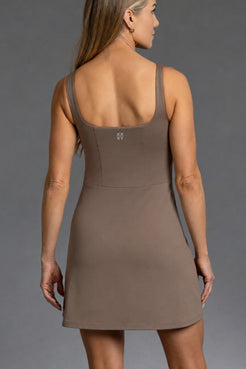 A woman shown from the back wearing a sleeveless, scoop-neck mini dress in a solid taupe-brown color, photographed against a neutral gray studio background. || mocha
