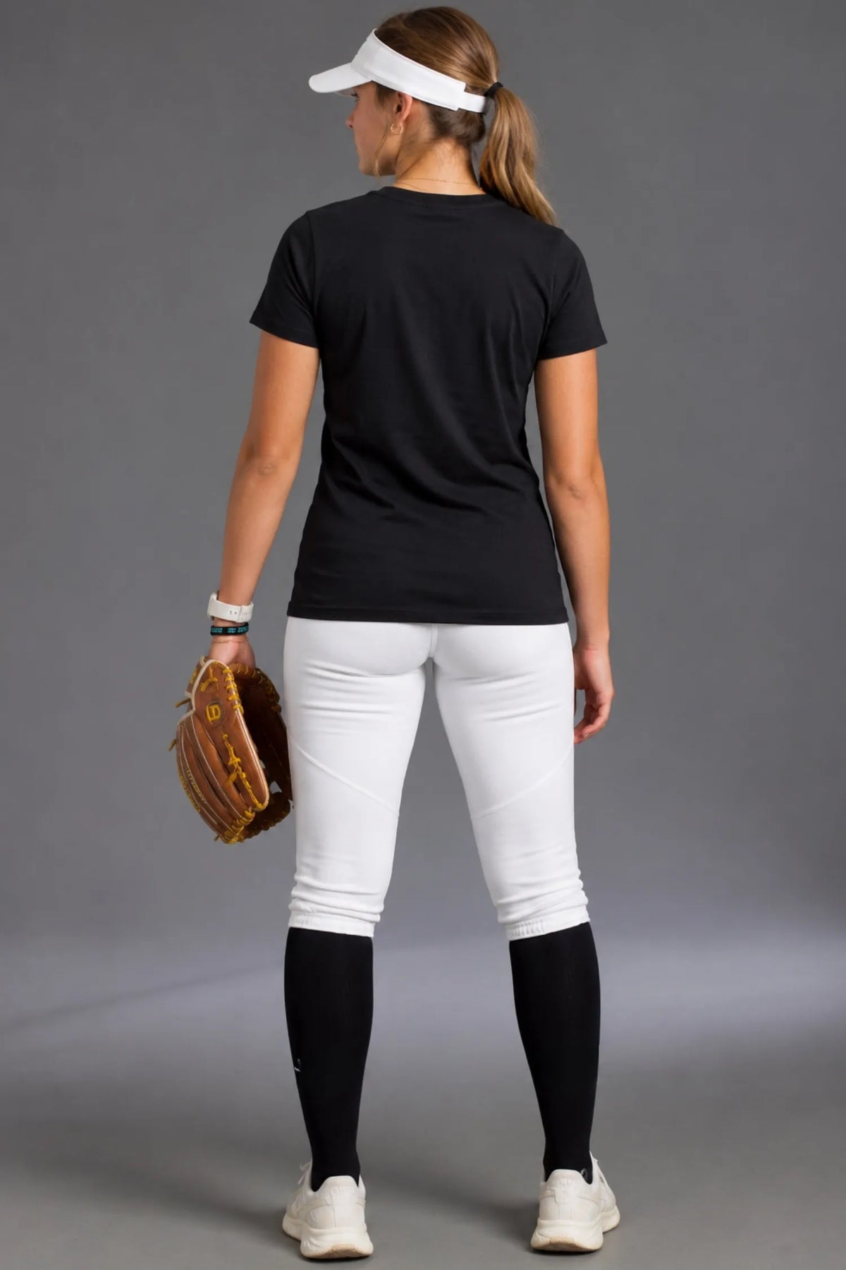 Rear view of a female softball player wearing a black XX-XY Athletics T-shirt with a blank back, white softball pants, black knee-high socks, and a white visor while holding a brown softball glove.