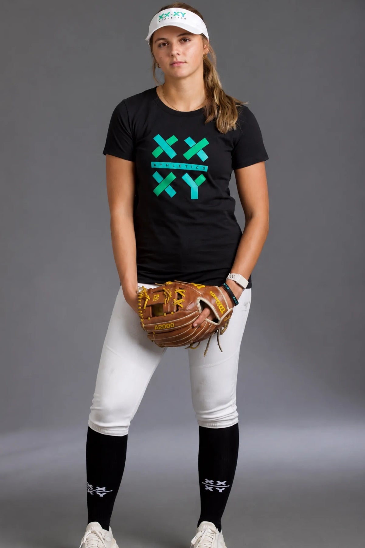 Female softball player wearing a black XX-XY Athletics graphic tee, white softball pants, black knee-high socks, and a white visor while holding a softball glove against a gray studio background.
