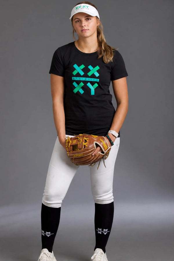 Female softball player wearing a black XX-XY Athletics graphic tee, white softball pants, black knee-high socks, and a white visor while holding a softball glove against a gray studio background. || Black