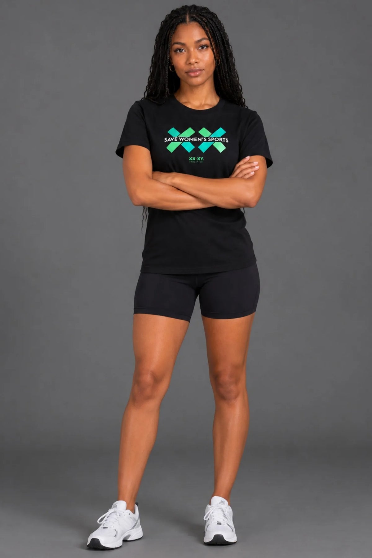 Female model wearing a black Save Women's Sports graphic tee with green and teal logo, paired with black biker shorts against a gray studio background