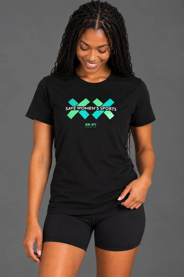 Female model wearing a black Save Women's Sports graphic tee with green and teal logo, paired with black biker shorts against a gray studio background || Black
