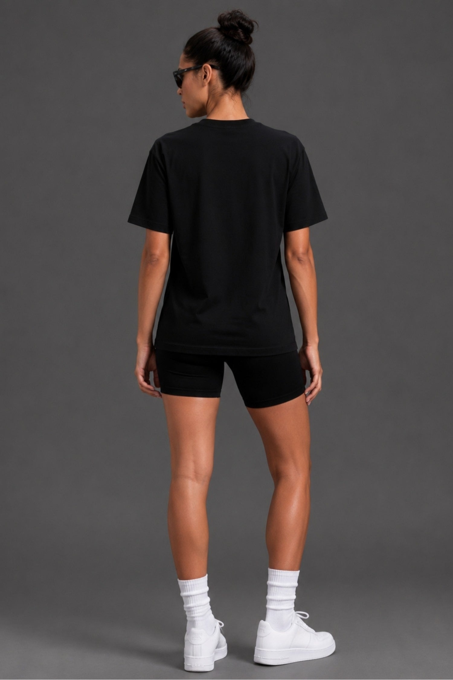 A woman modeling a black short-sleeve t-shirt  paired with black shorts, white socks, and white sneakers, set against a solid gray studio background.