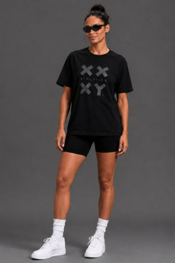 A woman modeling a black short-sleeve t-shirt with a gray XXYY logo graphic on the chest, paired with black shorts, white socks, and white sneakers, set against a solid gray studio background. || Black Tonal