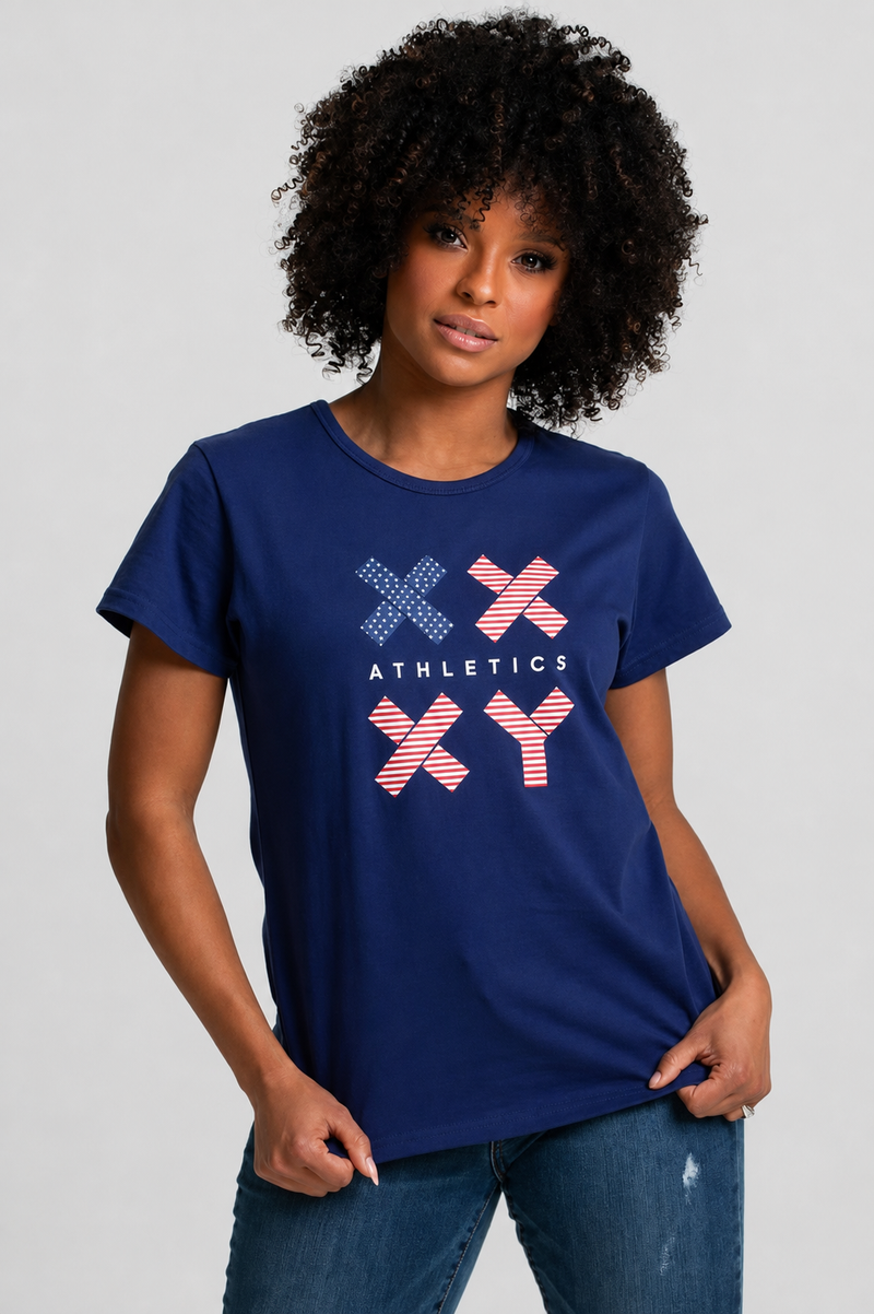 A woman with curly black hair wearing a navy blue short-sleeve t-shirt with a red, white, and blue graphic on the front, paired with blue jeans, against a light gray studio background. || Navy