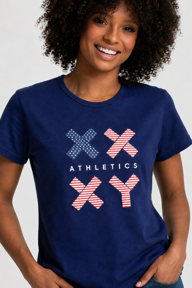 A woman with curly black hair wearing a navy blue short-sleeve t-shirt with a red, white, and blue graphic on the front, paired with blue jeans, against a light gray studio background. || Navy