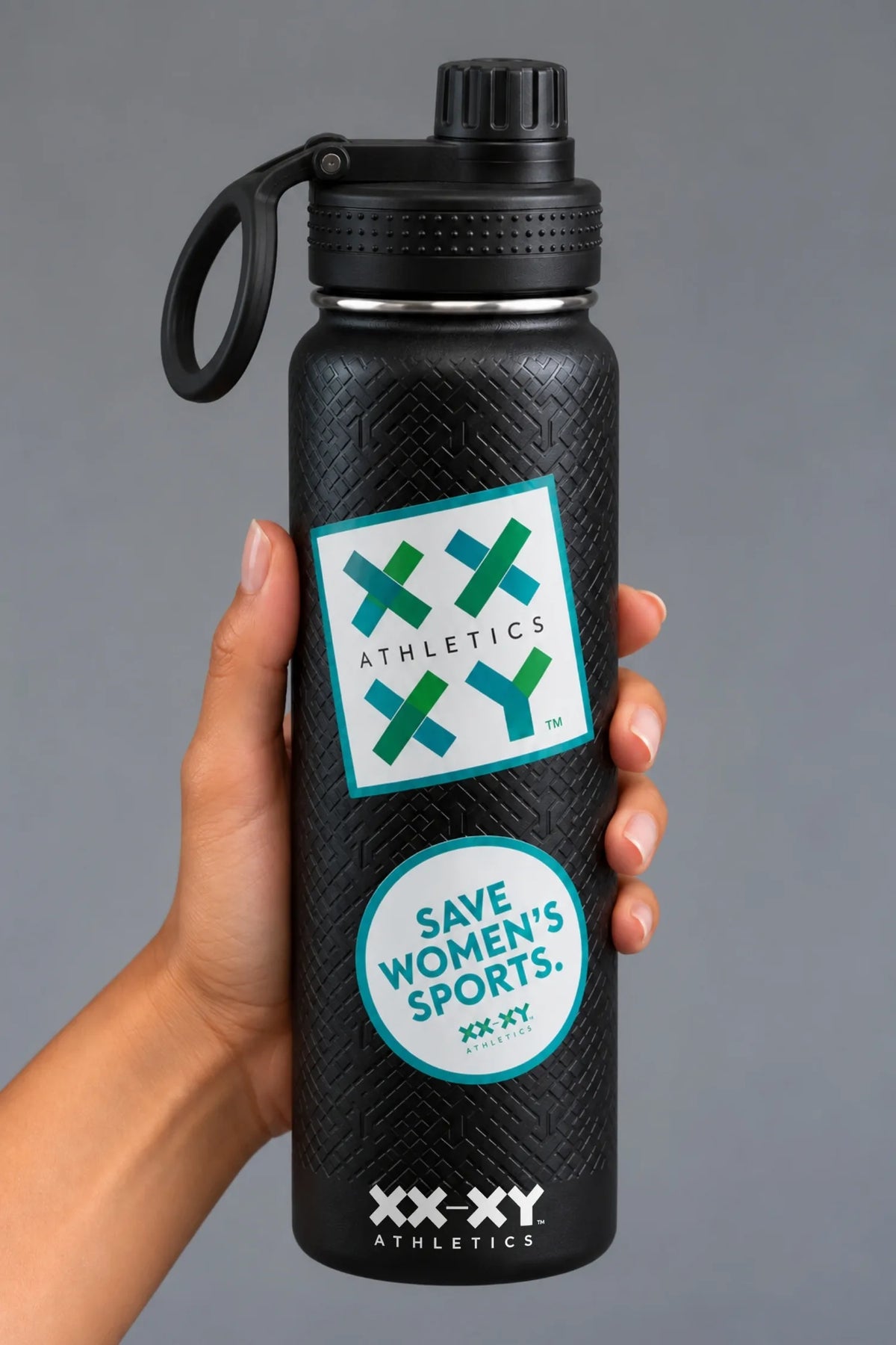 Hand holding black XX-XY Athletics water bottle with Athletics logo and Save Women’s Sports stickers on gray background || Black