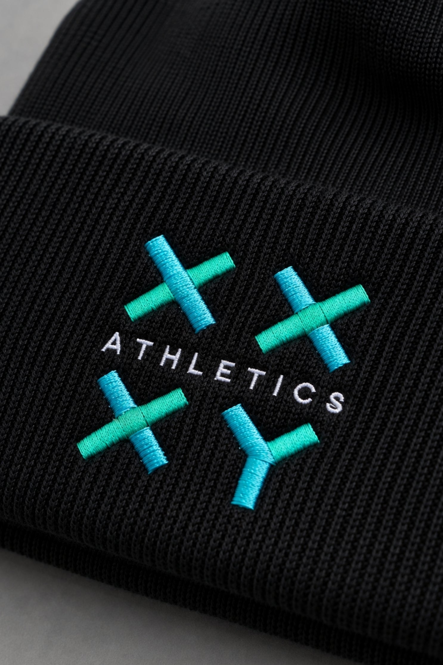 Close-up of black ribbed knit beanie featuring embroidered teal and blue XX-XY Athletics logo. || Black