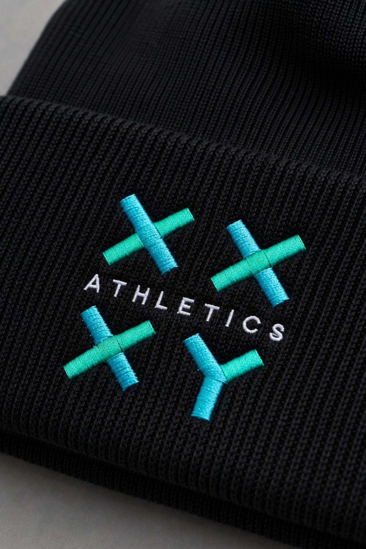 Close-up of black ribbed knit beanie featuring embroidered teal and blue XX-XY Athletics logo. || Black