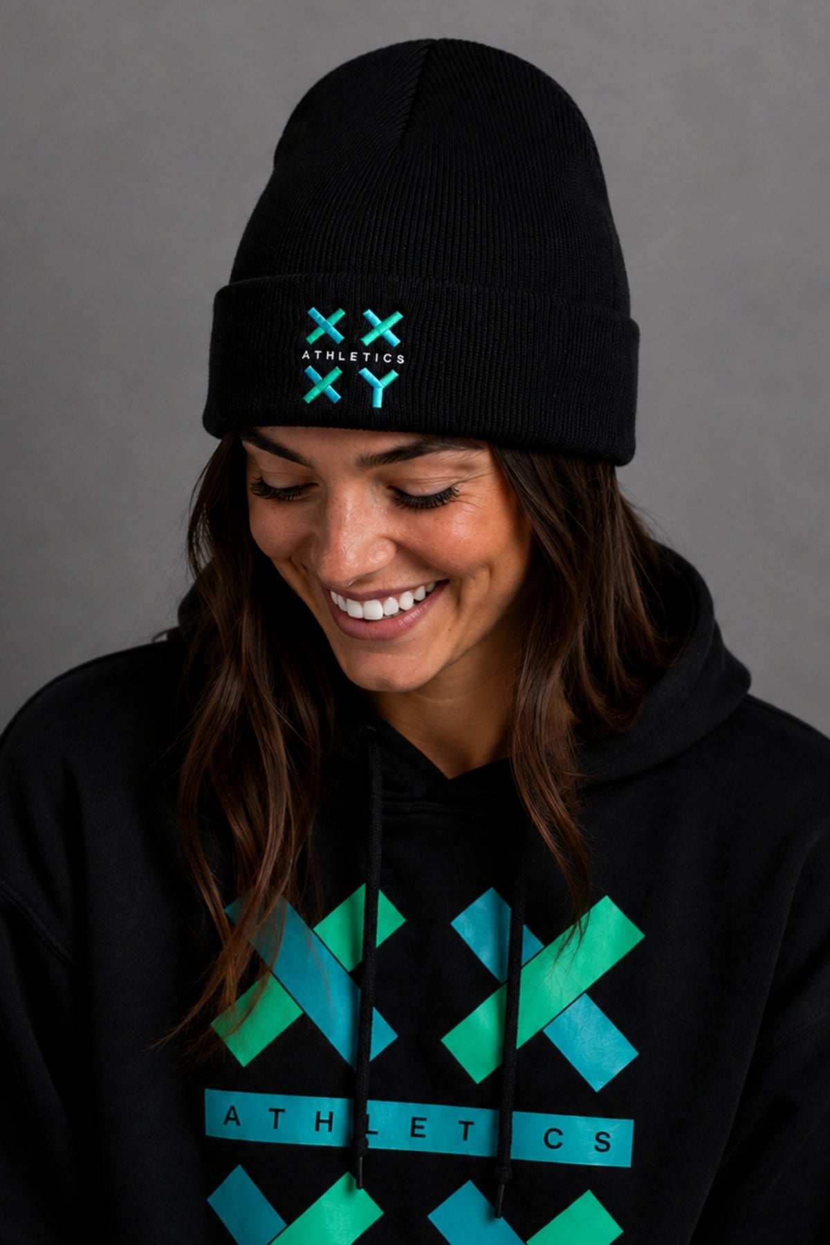 A woman wearing a black beanie and black hoodie with a teal and green geometric 'XX ATHLETICS' logo, shown against a plain gray background.