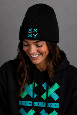 A woman wearing a black beanie and black hoodie with a teal and green geometric 'XX ATHLETICS' logo, shown against a plain gray background. || Black
