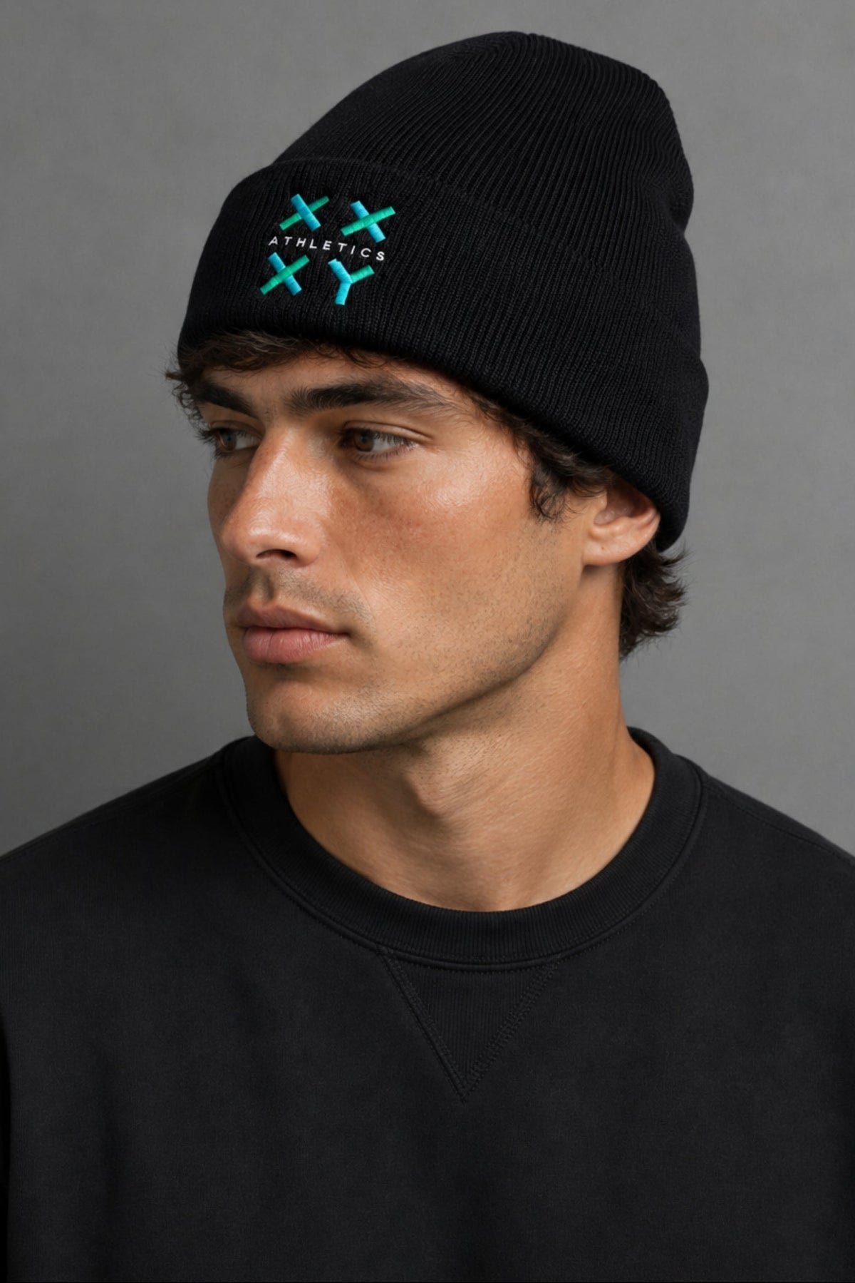 A man wearing a black knit beanie with a turquoise embroidered logo on the front, paired with a black crewneck sweatshirt against a neutral gray background.