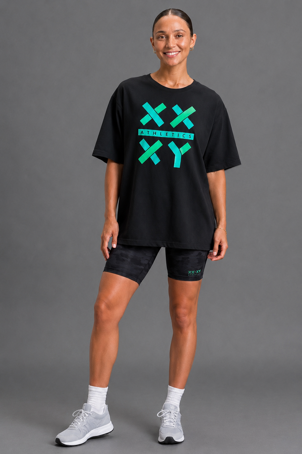 A woman modeling a black oversized graphic t-shirt with a bright green and teal 'XX XY' design on the front, paired with black athletic shorts, white socks, and gray sneakers against a solid gray studio background. || Black