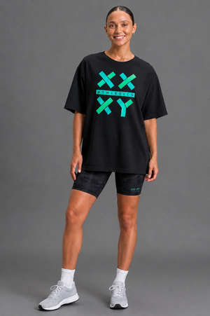 A woman modeling a black oversized graphic t-shirt with a bright green and teal 'XX XY' design on the front, paired with black athletic shorts, white socks, and gray sneakers against a solid gray studio background. || Black