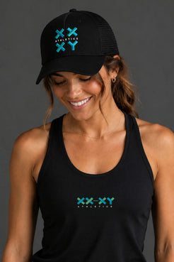 A woman wearing a black trucker hat and matching black tank top, both featuring a green and blue 'XXX-XY Athletics' logo on the front, against a solid dark gray background. || Black with Color Logo