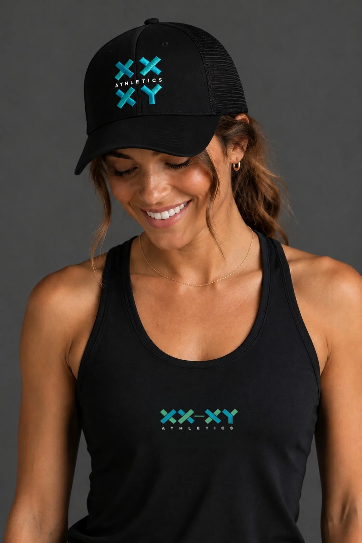 A woman wearing a black trucker hat and matching black tank top, both featuring a green and blue 'XXX-XY Athletics' logo on the front, against a solid dark gray background. || Black with Color Logo