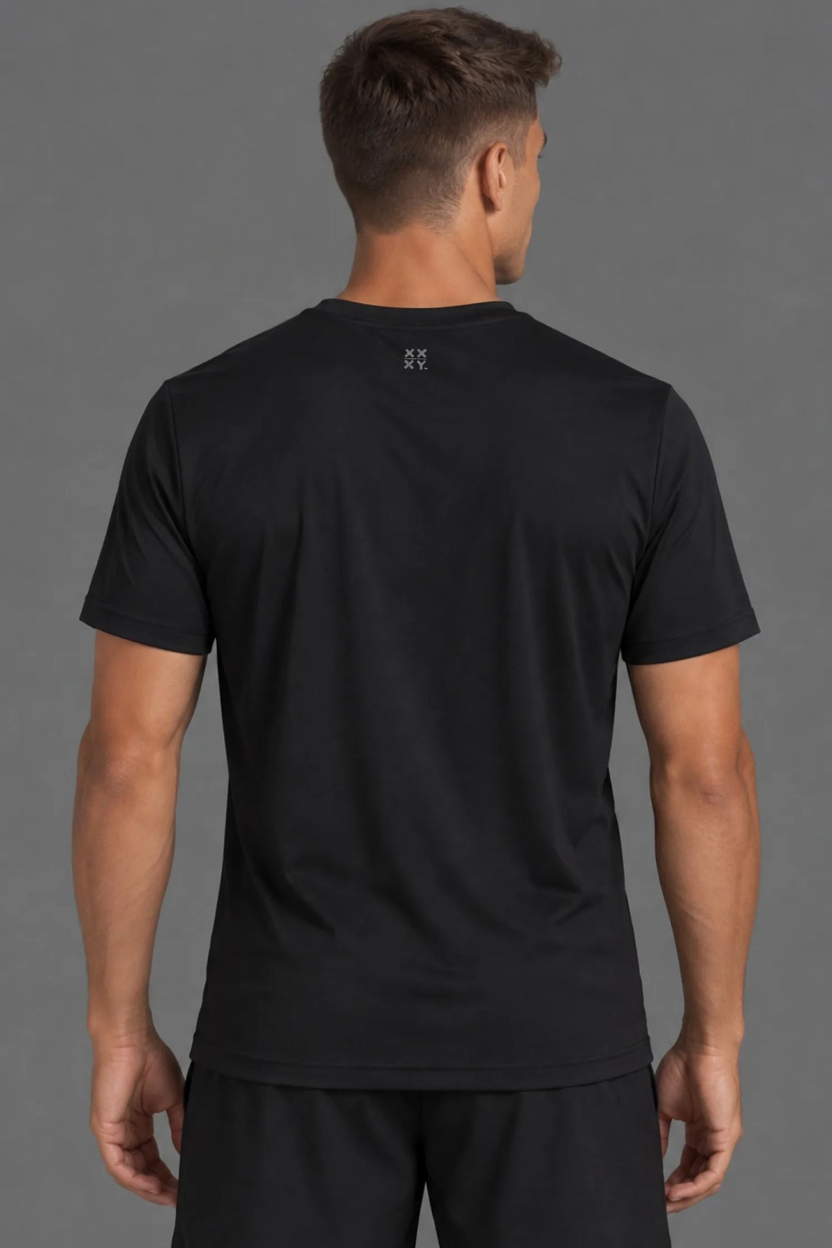 A man viewed from the back wearing a black short-sleeve athletic t-shirt with a small white logo centered below the collar, paired with black shorts, against a solid gray studio background.