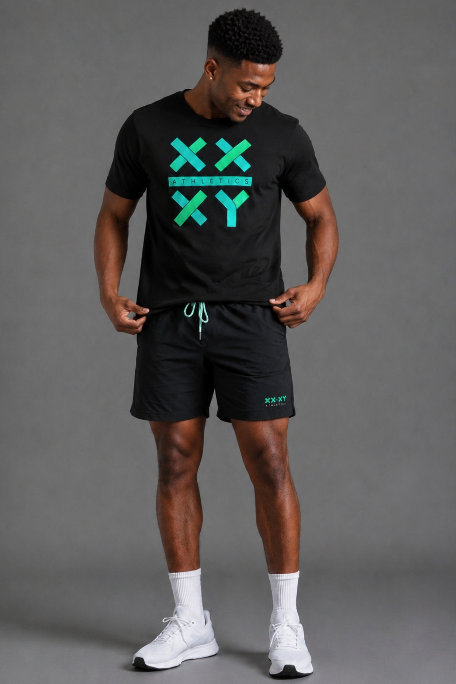 A man modeling a black athletic t-shirt and matching shorts set with a bright green XXYY graphic on the front, paired with white socks and sneakers, against a solid gray studio background. || Black