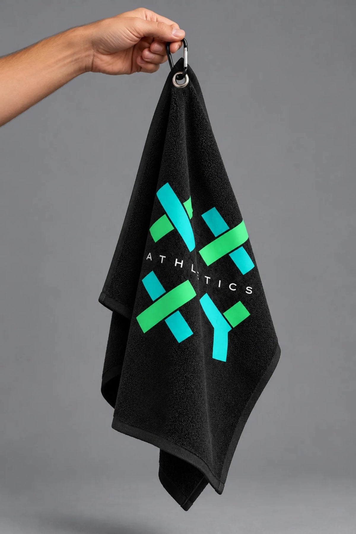 Hand holding a black XX-XY Athletics golf towel by its carabiner, showcasing the turquoise and green logo against a gray background || Black