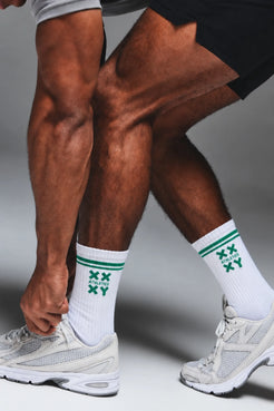 Male athlete tying his shoes while wearing white XX-XY Athletics crew socks with green stripes and logo detailing. || White