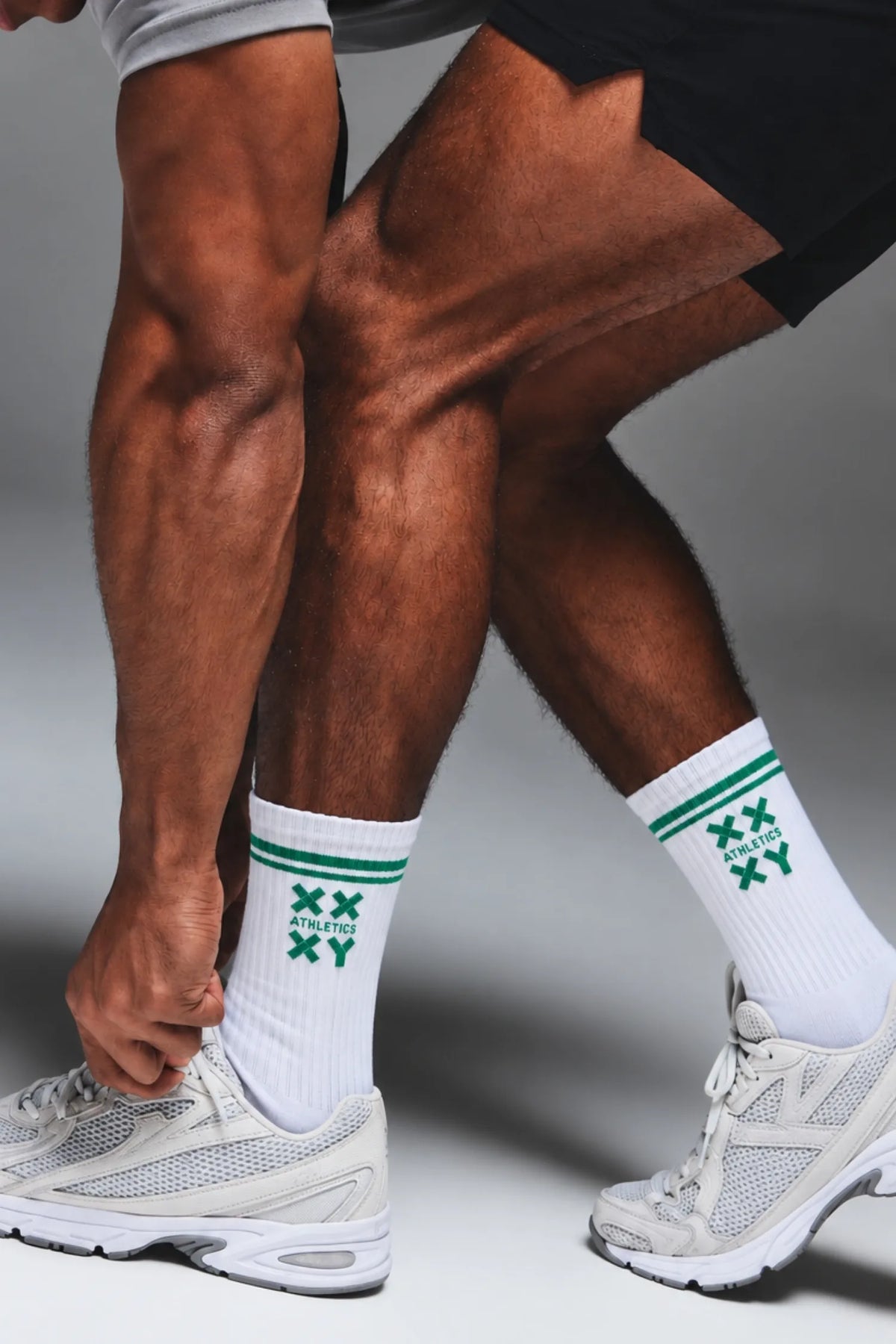 Male athlete tying his shoes while wearing white XX-XY Athletics crew socks with green stripes and logo detailing. || White