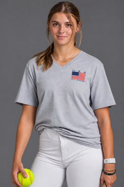Female softball player wearing a gray USA XX-XY Athletics V-neck T-shirt and white softball pants against a gray studio background. || Light Heather Gray