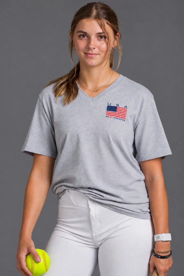 Female softball player wearing a gray USA XX-XY Athletics V-neck T-shirt and white softball pants against a gray studio background. || Light Heather Gray