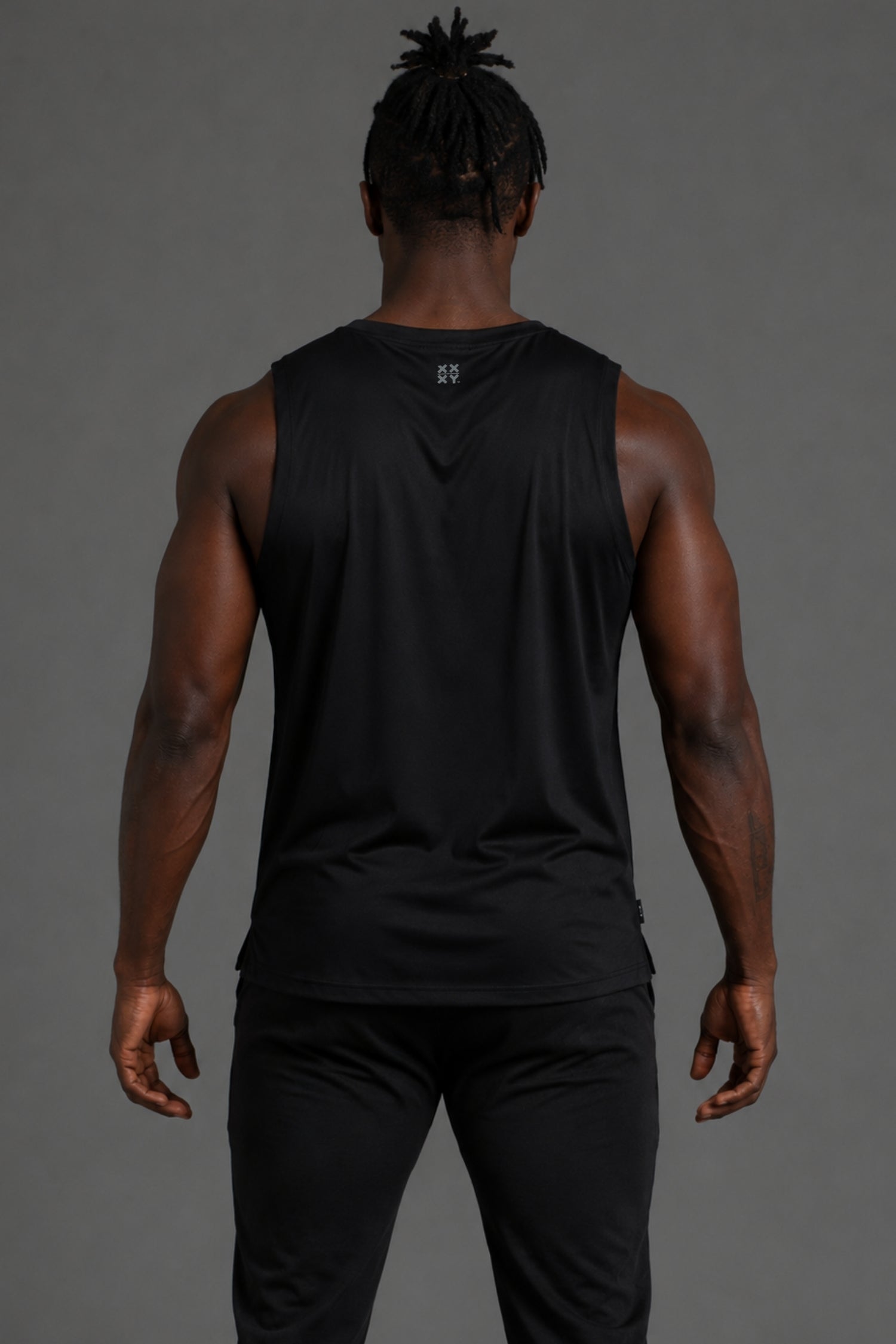A man viewed from behind wearing a black sleeveless athletic tank top and black pants against a solid gray studio background.