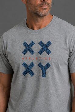 A close-up of a man wearing a heather gray t-shirt with a blue patterned XX and YY graphic and the word 'ATHLETICS' printed in red on the chest, set against a solid gray background. || Gray Camo