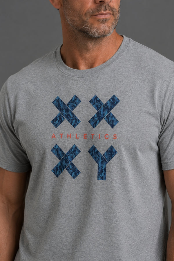 A close-up of a man wearing a heather gray t-shirt with a blue patterned XX and YY graphic and the word 'ATHLETICS' printed in red on the chest, set against a solid gray background. || Gray Camo
