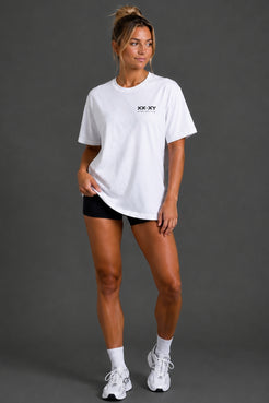 A woman modeling a white short-sleeve crewneck t-shirt with a small black 'XX.XY' logo on the chest, paired with black athletic shorts, white socks, and white sneakers, against a dark gray studio background. || White