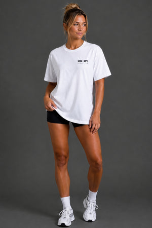 A woman modeling a white short-sleeve crewneck t-shirt with a small black 'XX.XY' logo on the chest, paired with black athletic shorts, white socks, and white sneakers, against a dark gray studio background. || White