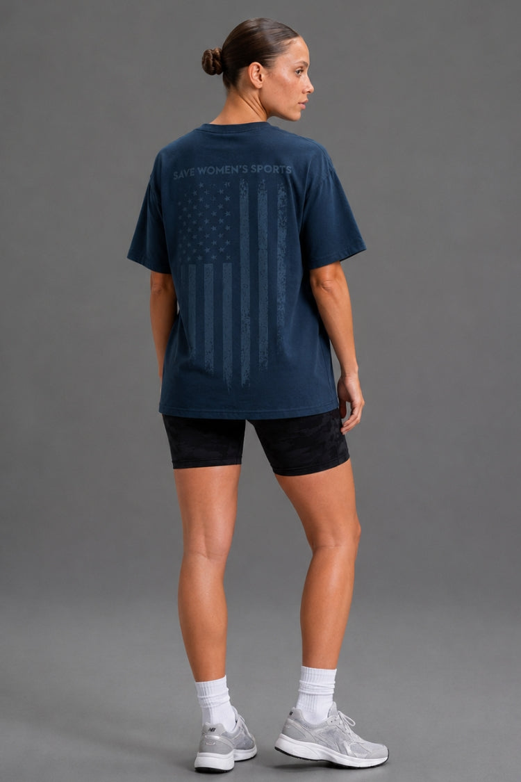 A woman shown from the back wearing a navy blue athletic T-shirt with a faded American flag graphic and 'WOMEN'S SPORTS' text, paired with black shorts, white socks, and gray sneakers against a gray studio background.