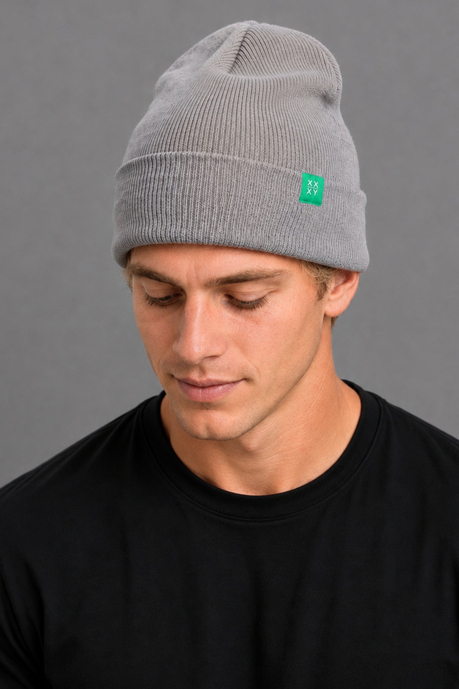 A man wearing a light gray ribbed knit beanie with a small green square logo patch on the side, paired with a black t-shirt against a solid gray background.