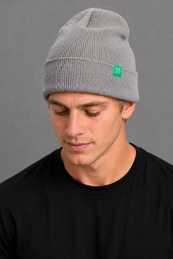 A man wearing a light gray ribbed knit beanie with a small green square logo patch on the side, paired with a black t-shirt against a solid gray background. || Light Heather Gray