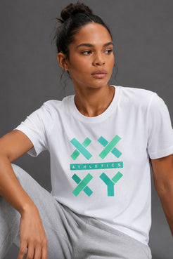 A woman wearing a white crewneck t-shirt with a green and teal 'XX ATHLETICS YY' graphic on the front, paired with light gray sweatpants, set against a solid dark gray studio background. || White