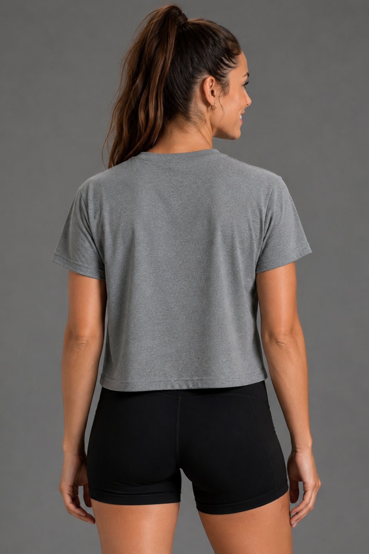 A woman shown from the back wearing a heather gray short-sleeve crop top and black athletic shorts against a solid gray studio background. || Heather Gray