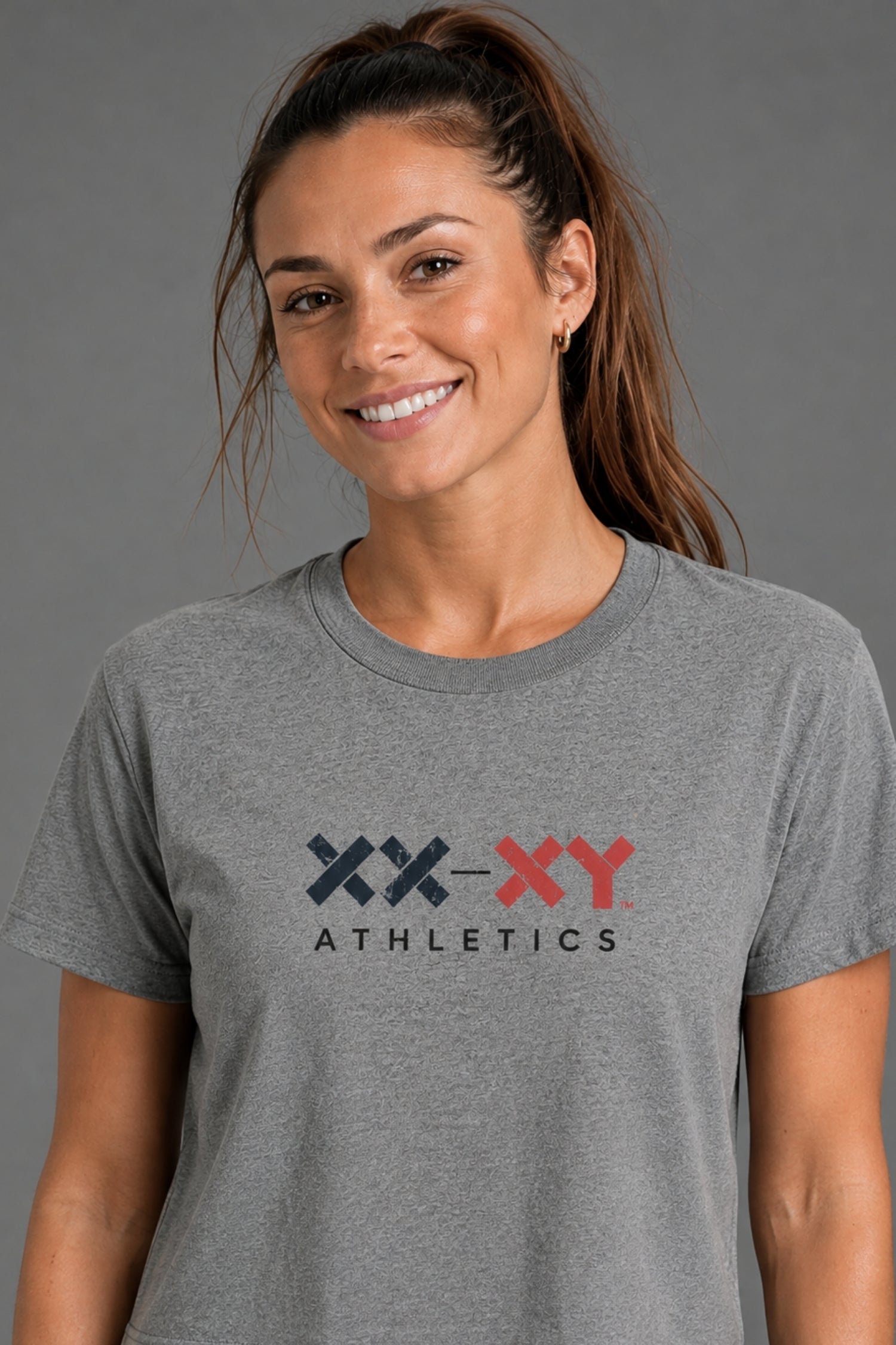 A woman modeling a heather gray cropped t-shirt with a 'XX-XY ATHLETICS' logo on the chest against a solid gray studio background.