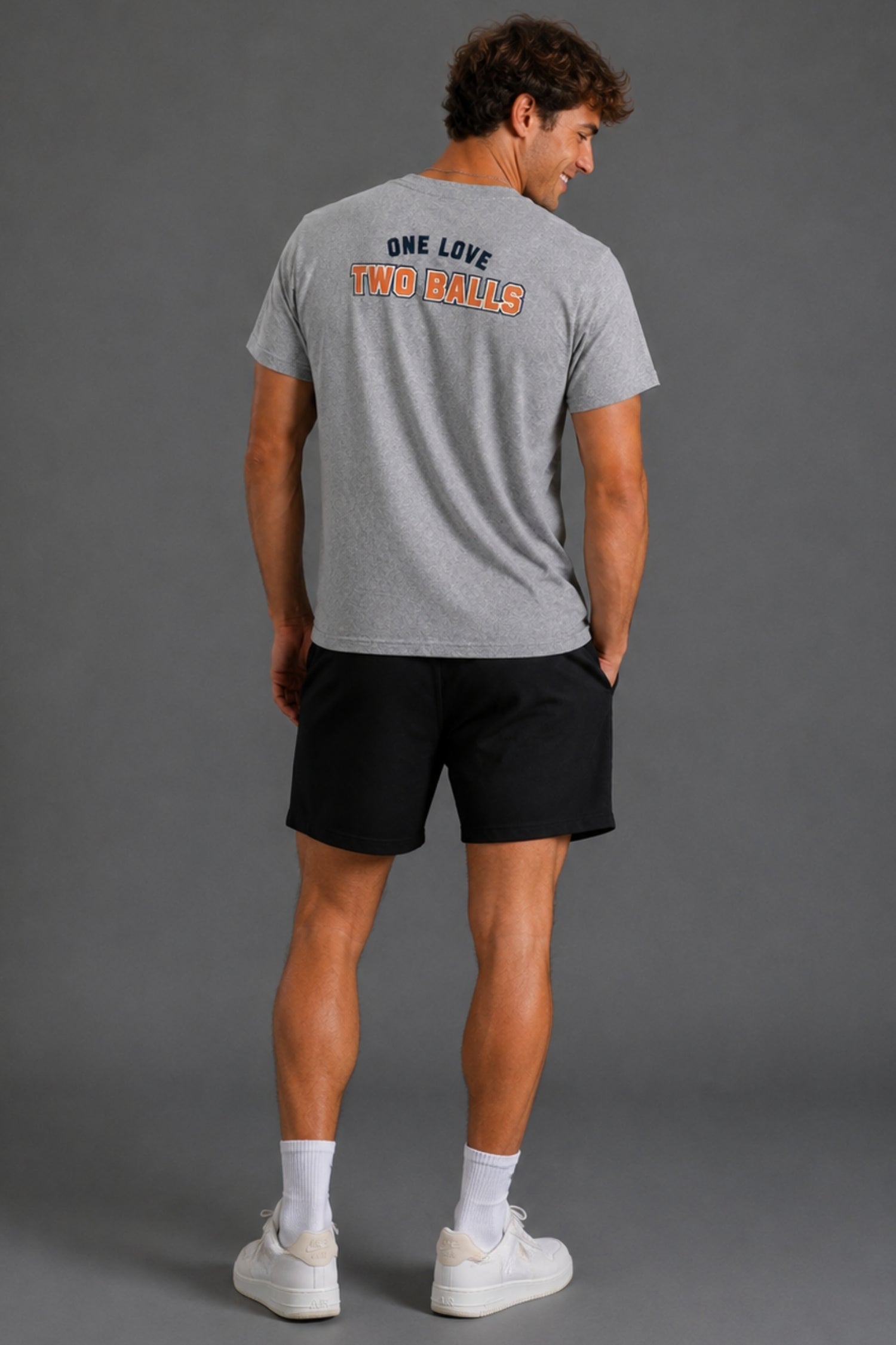 A man wearing a heather gray t-shirt with the text 'ONE LOVE TWO BALLS' printed on the back, paired with black athletic shorts, white socks, and white sneakers, photographed from behind against a solid gray studio background.