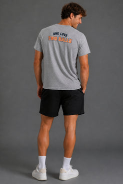 A man wearing a heather gray t-shirt with the text 'ONE LOVE TWO BALLS' printed on the back, paired with black athletic shorts, white socks, and white sneakers, photographed from behind against a solid gray studio background. || Heather Gray