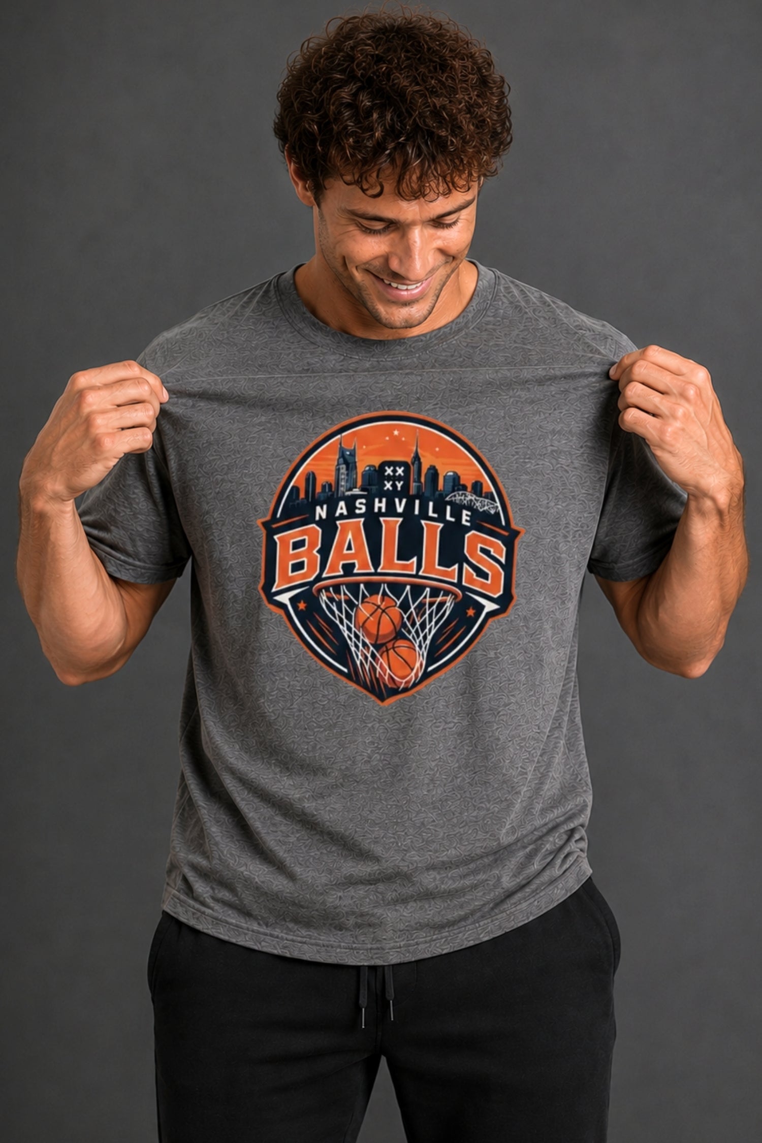 A man wearing a heather gray t-shirt with a 'Nashville Balls' basketball graphic on the front, holding the fabric to show its stretch against a dark gray studio background. || Dark Heather Gray