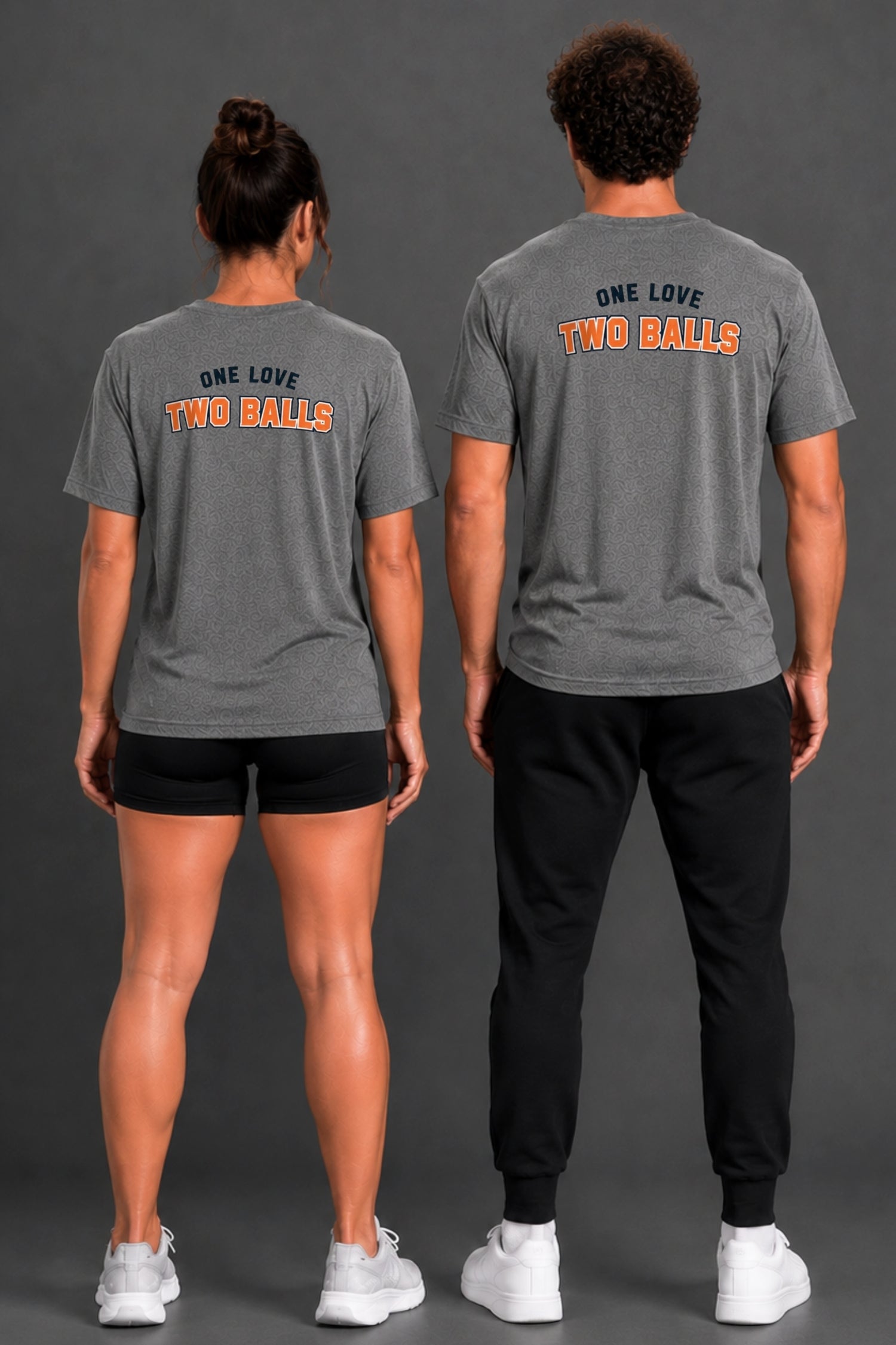 Two models shown from the back wearing matching dark gray t-shirts with the text 'ONE LOVE TWO BALLS' printed in navy blue and orange. The model on the left wears black athletic shorts and white sneakers, while the model on the right wears black jogger pants and white sneakers, set against a dark gray studio background. || Dark Heather Gray