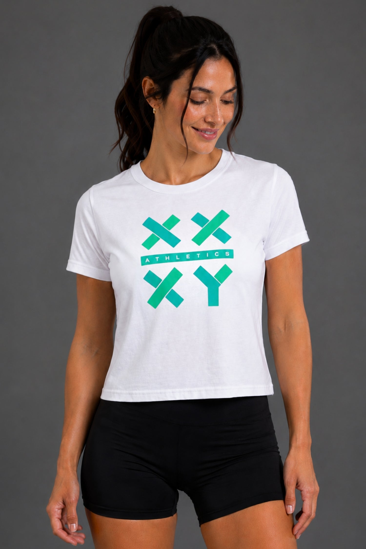 A woman wearing a white short-sleeve athletic crop top with a teal 'XXY ATHLETICS' graphic on the front, paired with black athletic shorts, against a solid gray studio background. || White