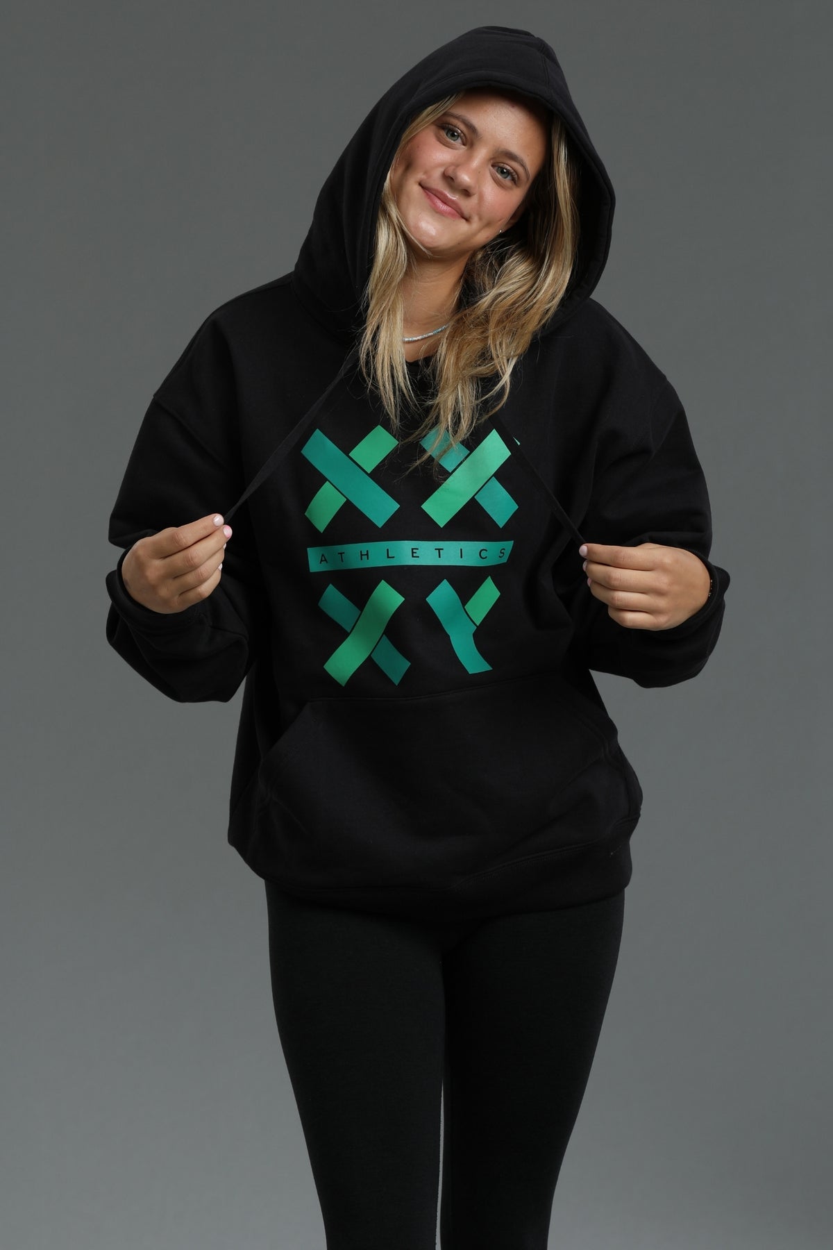 A woman wearing a black pullover hoodie with a green and teal graphic on the front that reads 'XX ATHLETIC', posed against a solid gray studio background. || Black