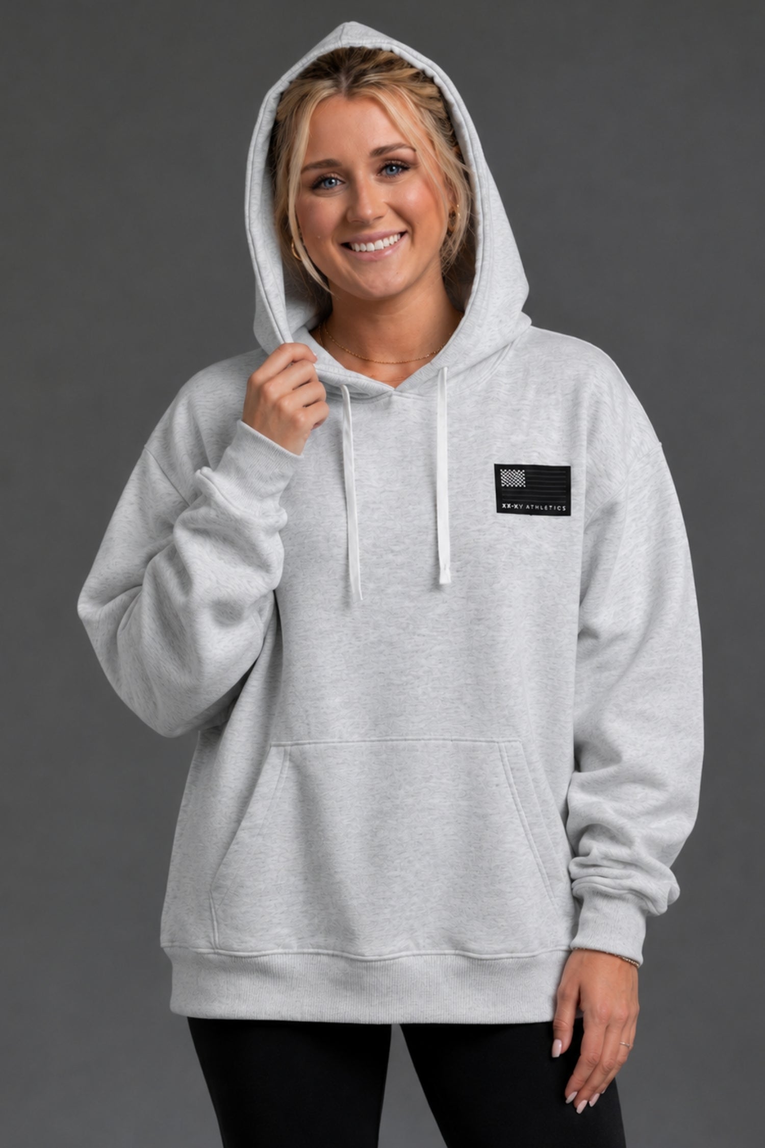 A woman wearing a light gray heather pullover hoodie with a small black rectangular logo patch on the left chest, posed against a solid dark gray studio background.