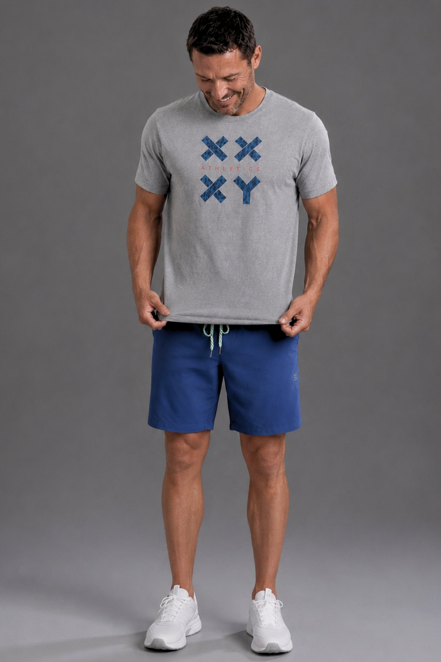 A man modeling a heather gray t-shirt with a blue 'XX XY' graphic on the chest, paired with blue athletic shorts and white sneakers, against a solid gray studio background. || Gray Camo
