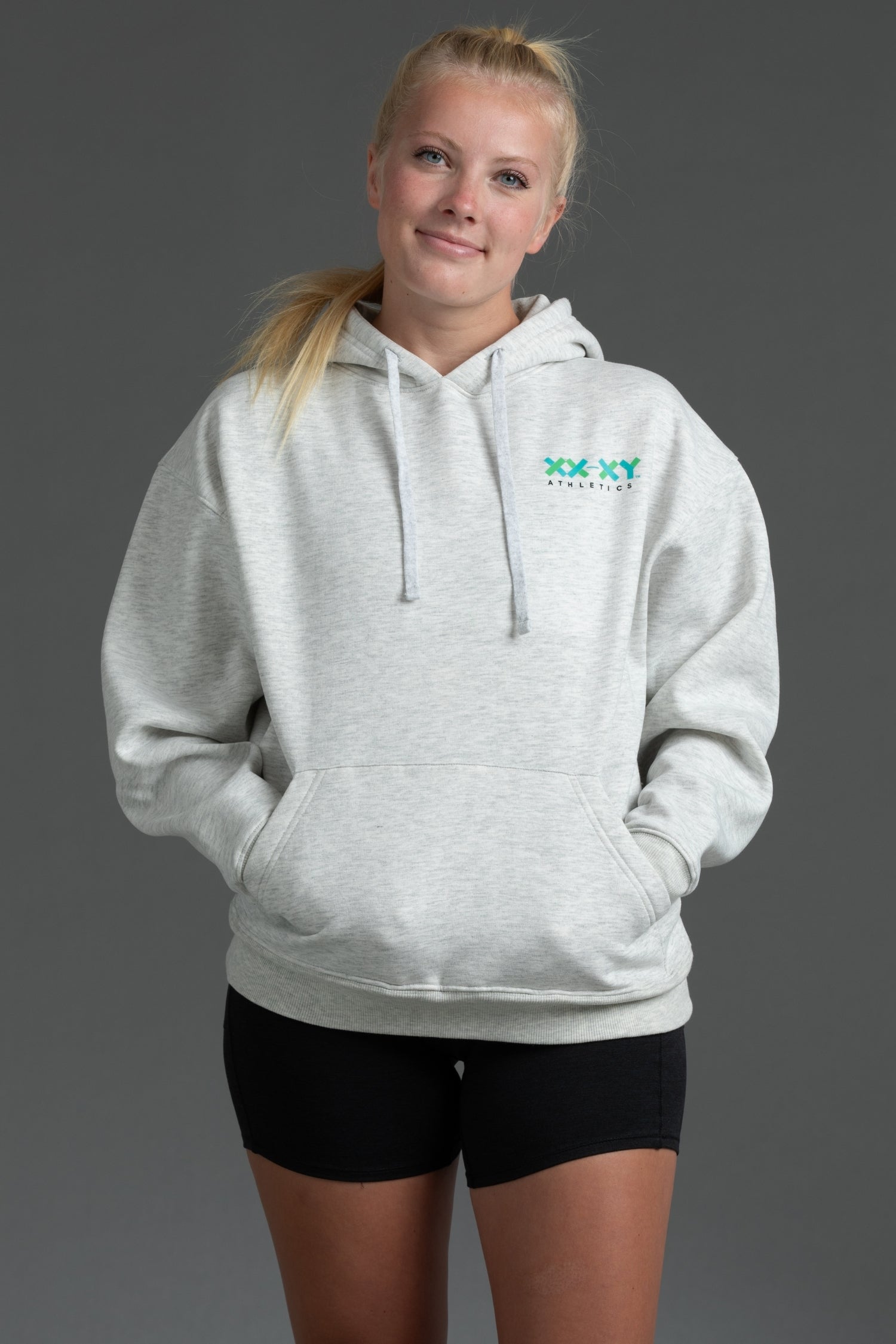 A woman modeling a light heather gray pullover hoodie with a green 'XXX YYYathletics' logo on the left chest, paired with black athletic shorts, against a solid dark gray studio background. || Light Heather Gray